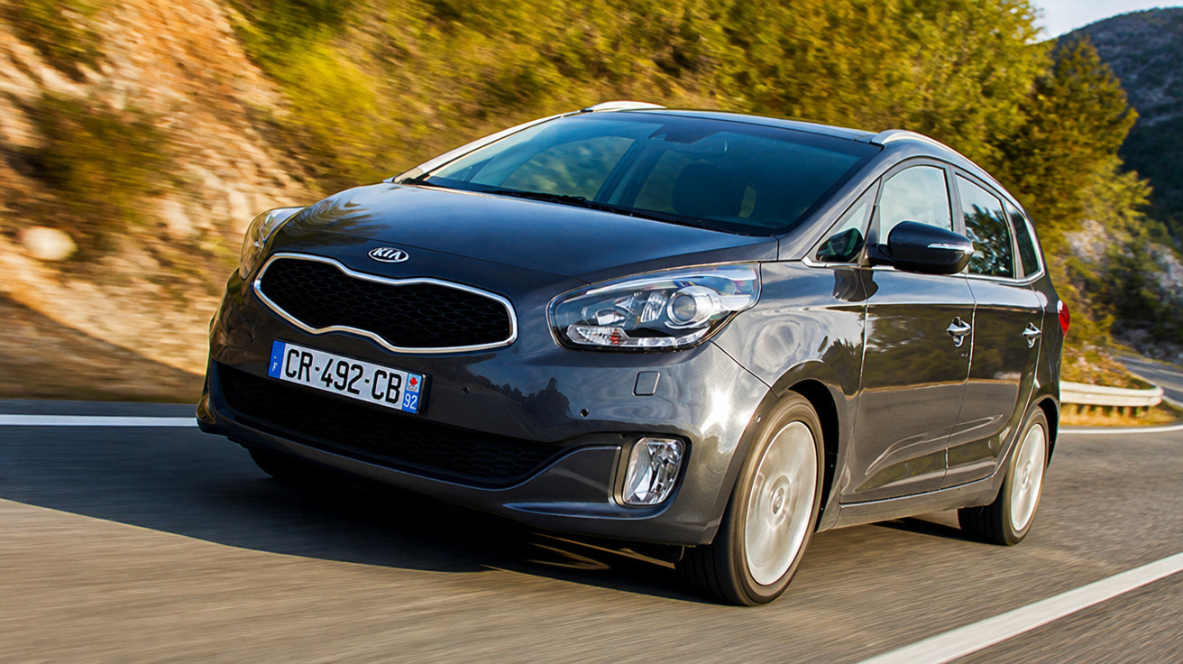 Carro Kia cinza escuro em movimento em estrada com vegetação e montanhas ao fundo.