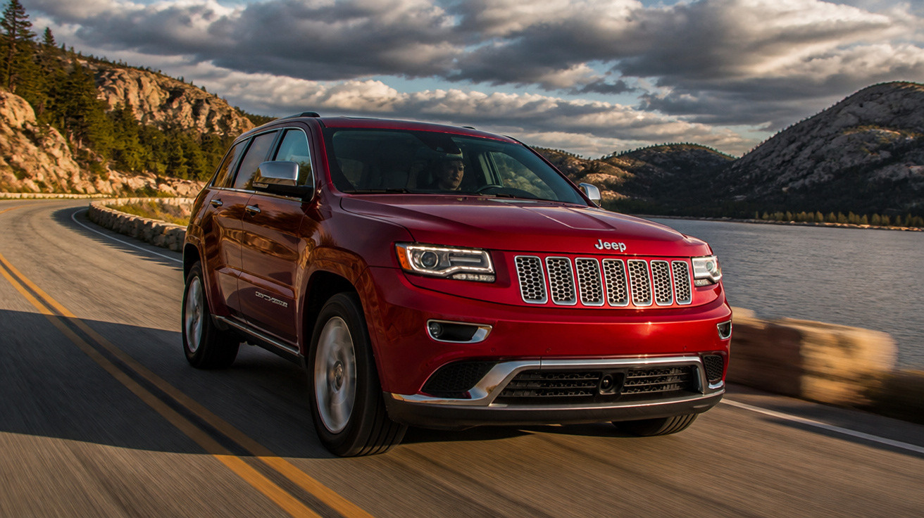 SUV vermelho da Jeep dirigindo em estrada curvada junto a lago e montanhas sob céu nublado.