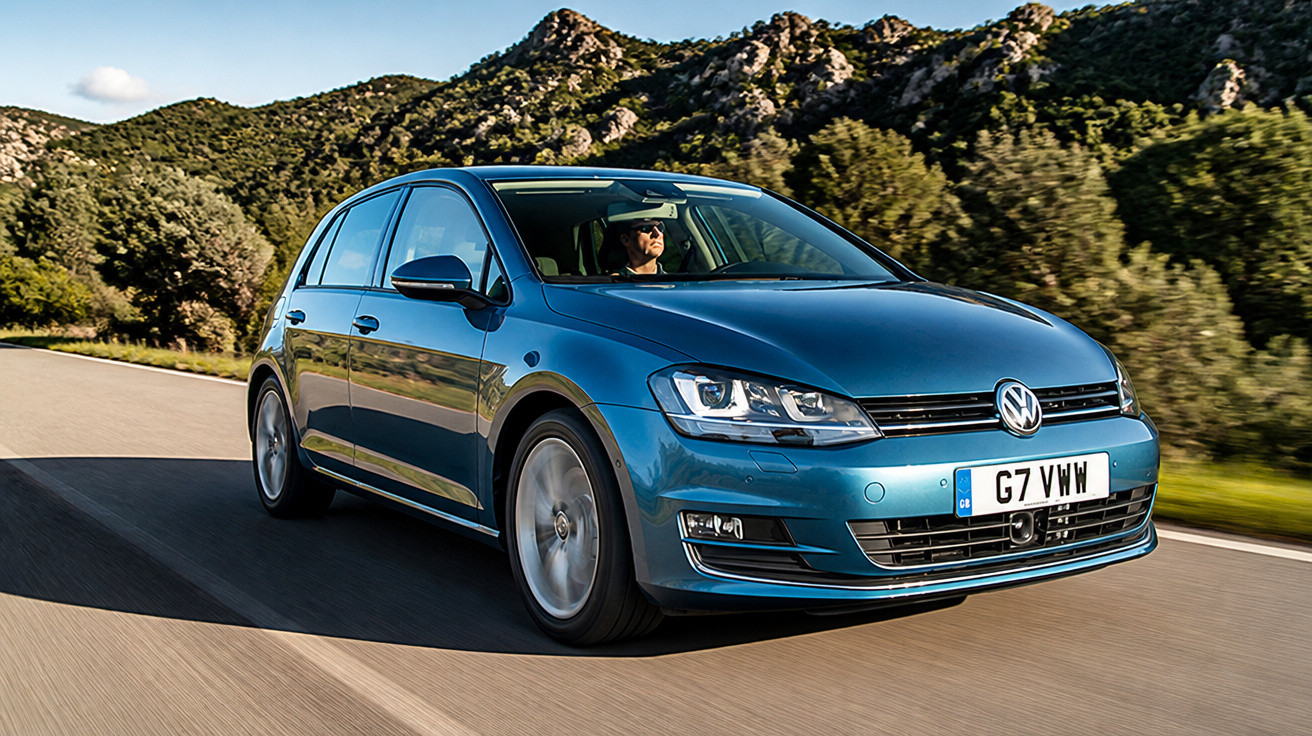 Carro Volkswagen azul em movimento em estrada rodeada por árvores e montanhas no fundo.