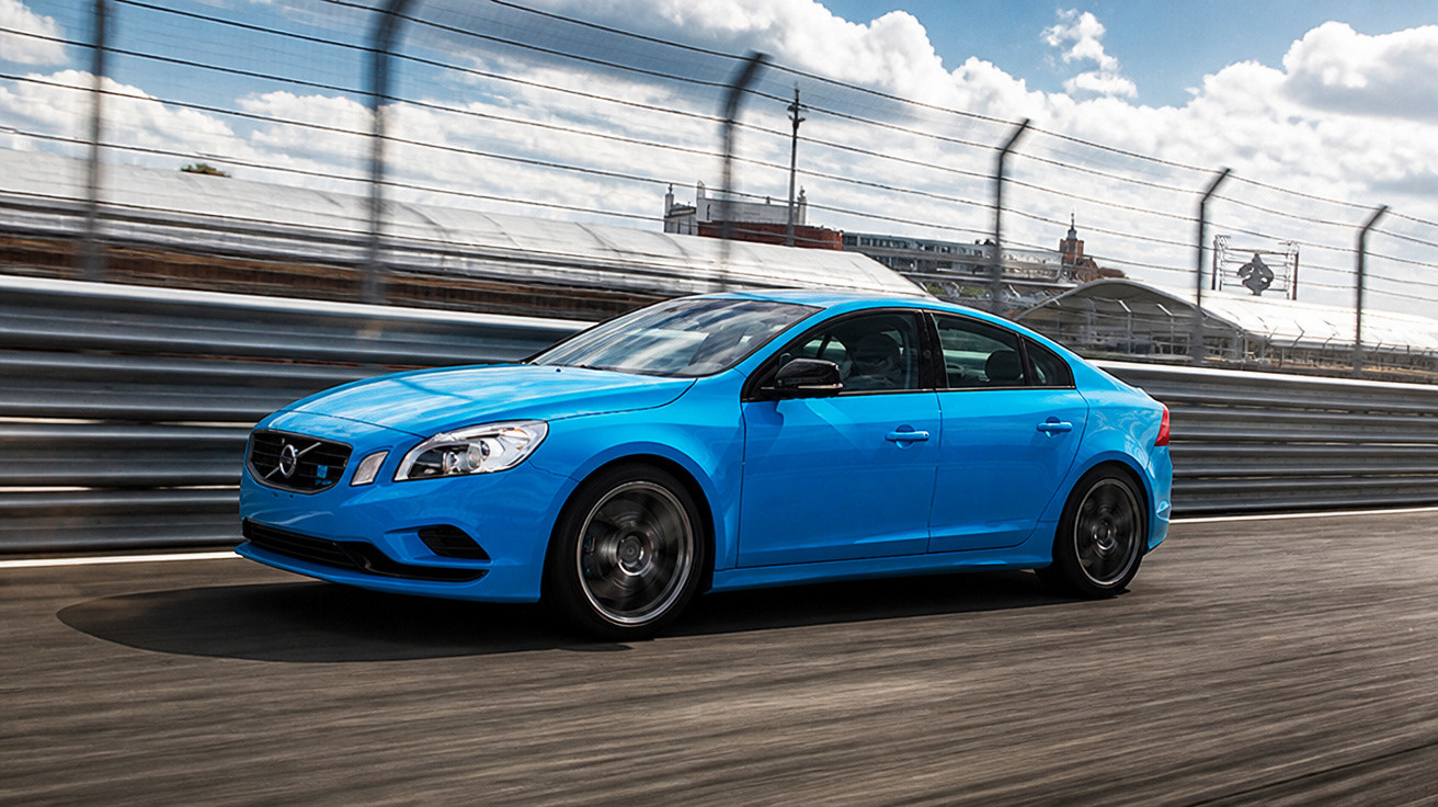 Carro sedan esportivo azul em pista de corrida com grade de proteção e céu parcialmente nublado ao fundo.