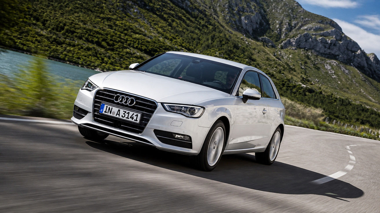 Carro Audi branco em movimento em estrada sinuosa perto de lago e montanhas.