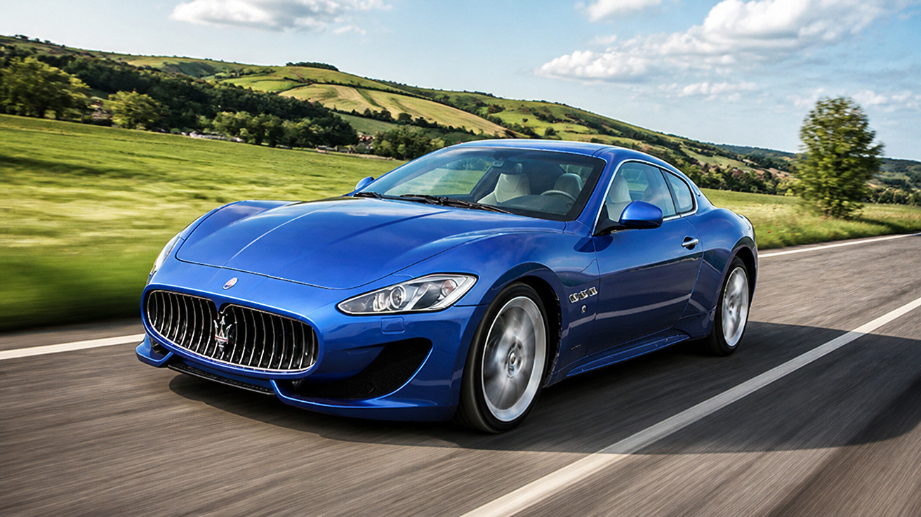 Carro esportivo azul modelo Maserati GranTurismo circulando em estrada cercada por campo verde e colinas.