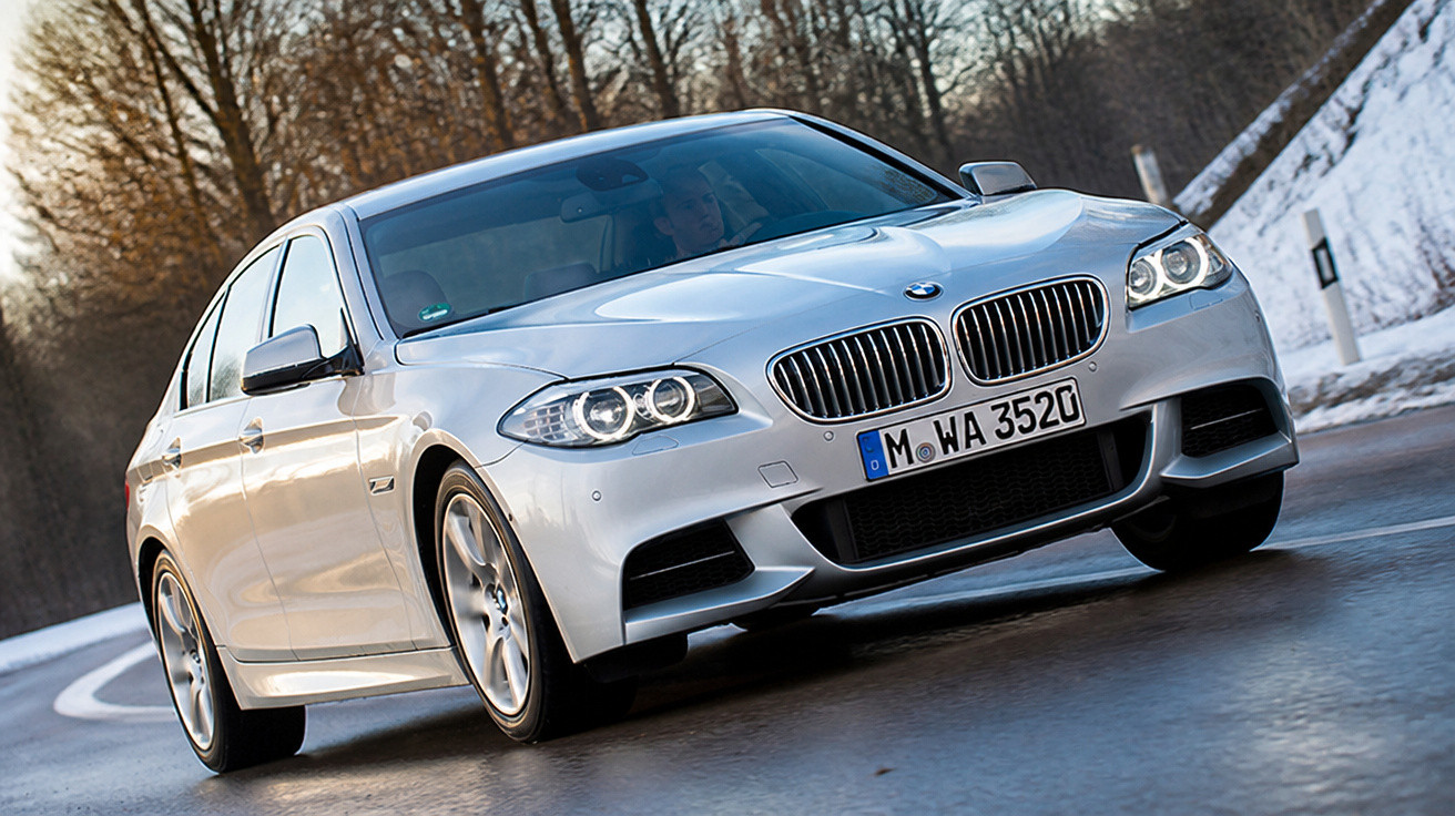 Carro sedã prata BMW em estrada molhada com árvores e neve ao fundo em dia nublado.