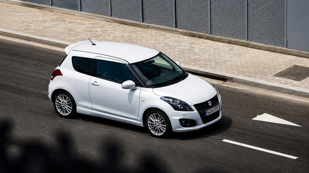Carro branco hatchback Suzuki Swift em uma rua asfaltada com calçada de pedras.