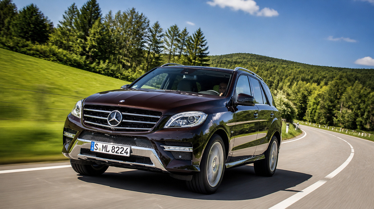 Carro Mercedes-Benz SUV marrom escuro em estrada sinuosa cercada por árvores e céu azul.