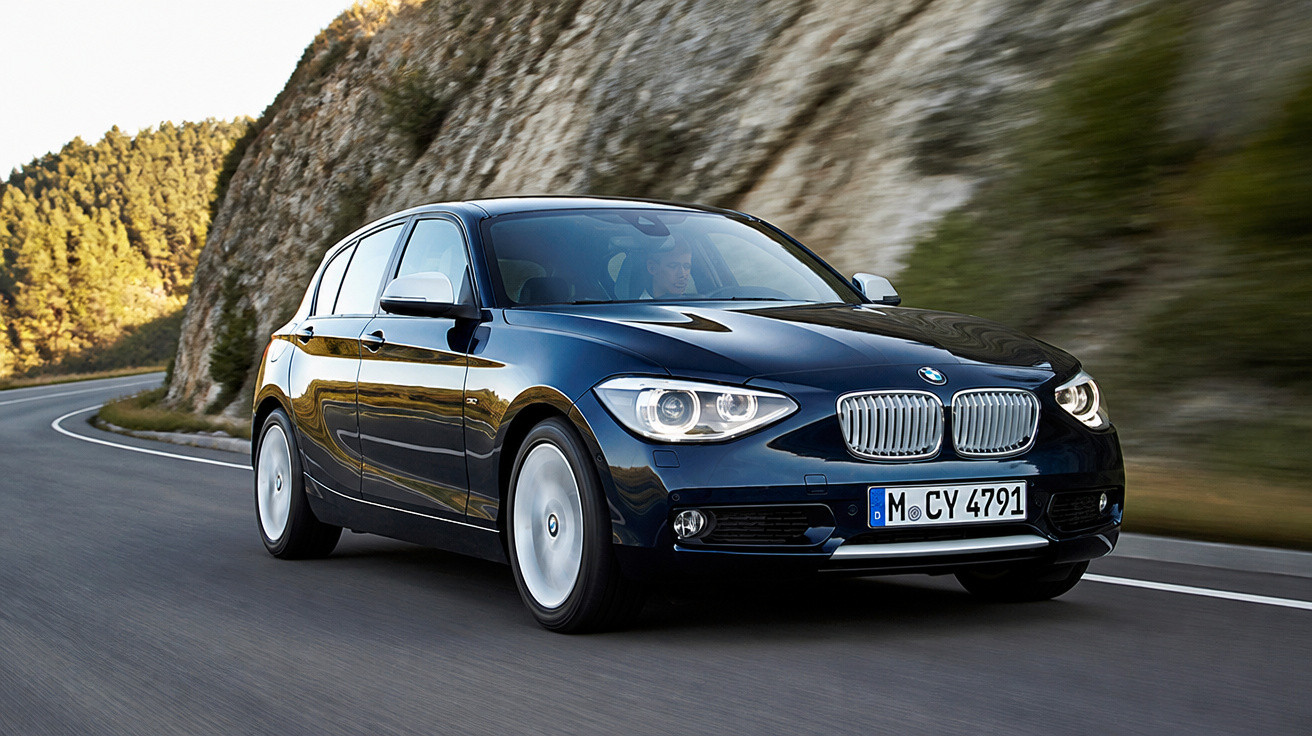 Carro preto BMW hatchback circulando em estrada curva com montanhas ao fundo.
