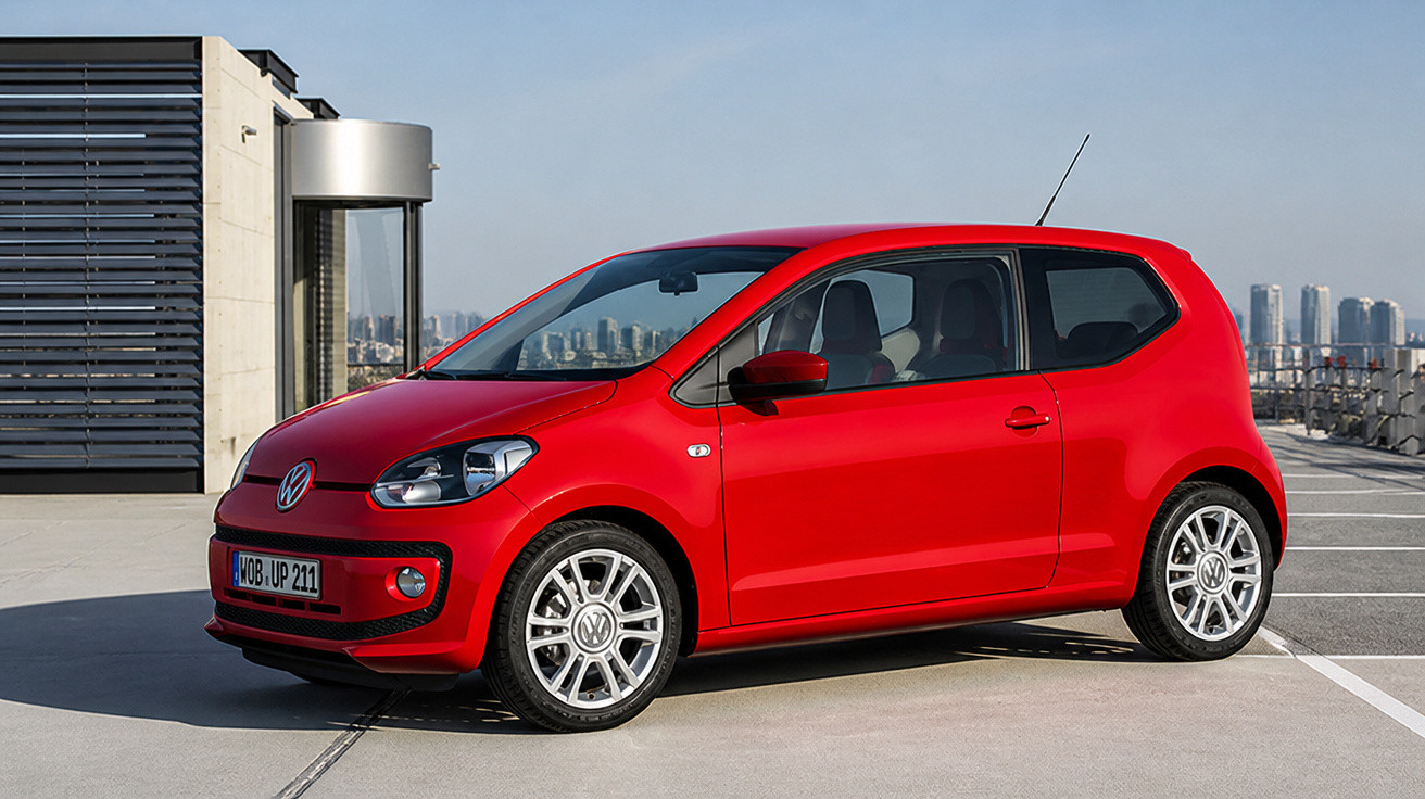 Carro vermelho Volkswagen compacto estacionado em área urbana com arranha-céus ao fundo.