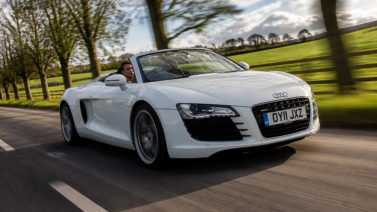 Carro conversível esportivo branco Audi R8 dirigindo em estrada com árvores e campos ao fundo.