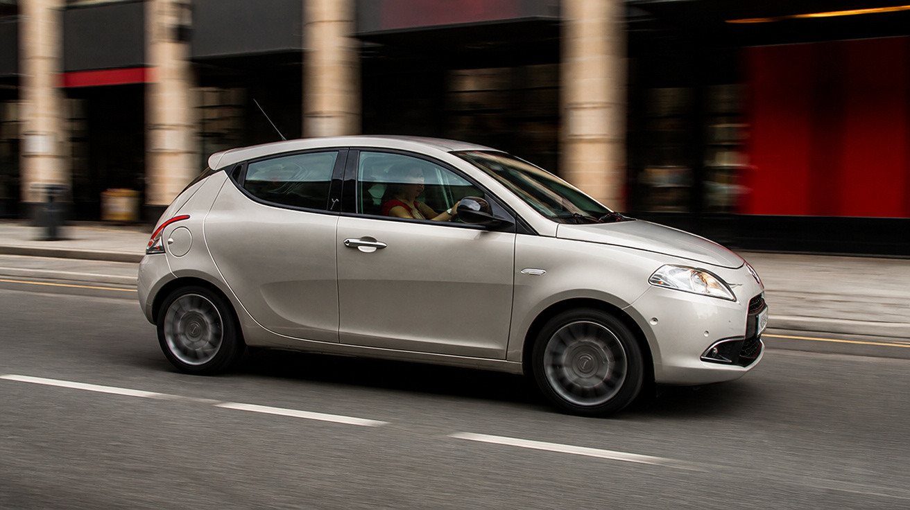 Carro prata hatchback em movimento em uma rua urbana com prédios ao fundo.