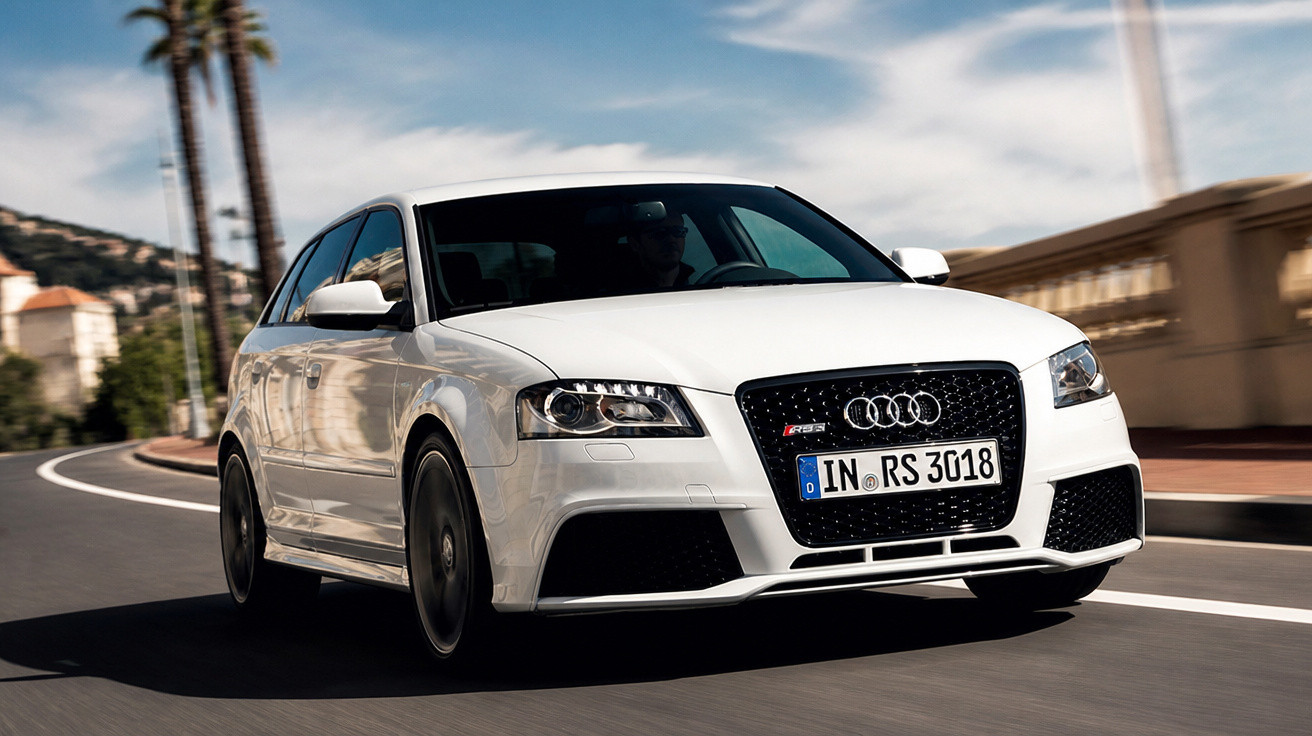 Carro Audi branco modelo RS3 Sportback em movimento em estrada curva com palmeiras ao fundo.