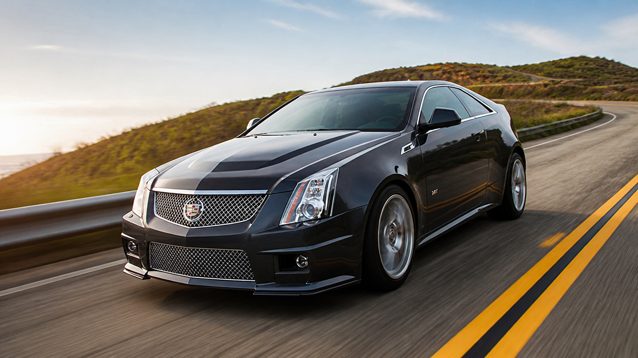 Carro esportivo Cadillac preto em estrada sinuosa ao entardecer com colinas ao fundo.